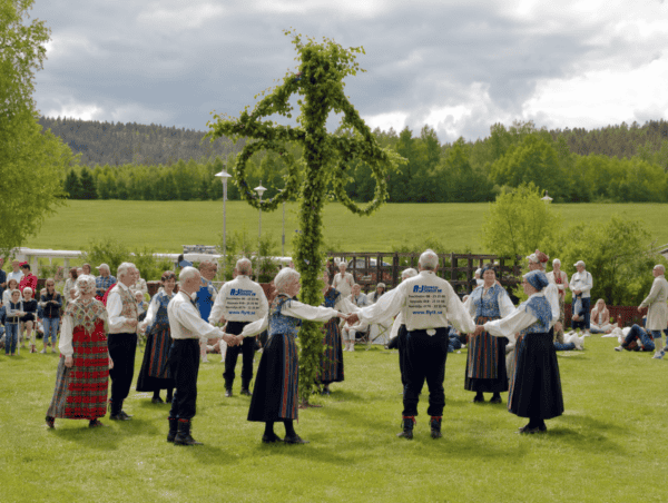 Folkdansare i traditionella kläder dansar runt en midsommarstång utomhus.