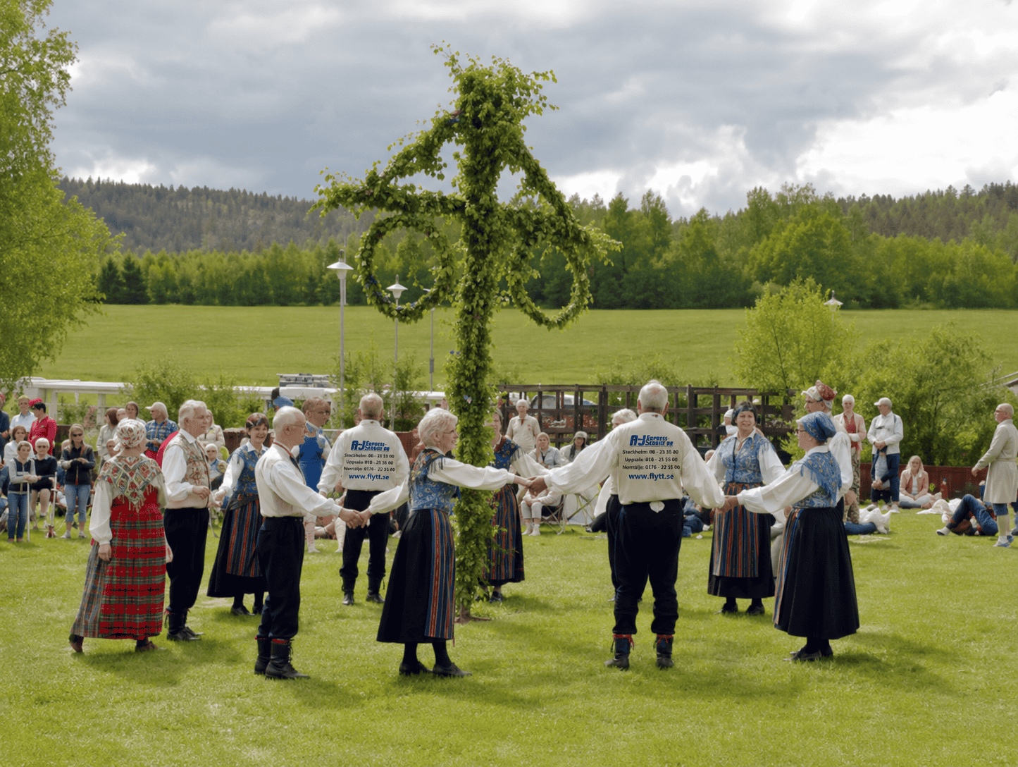 Folkdansare i traditionella kläder dansar runt en midsommarstång utomhus.
